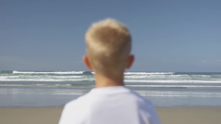 A blonde haired boy stands on the sand and watches the waves curl and break as they come to shore