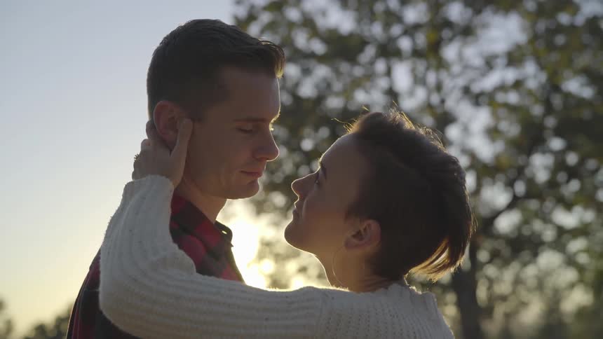 A teenage boy and a teenage girl stand outside in the sunset holding each other and looking at each other lovingly