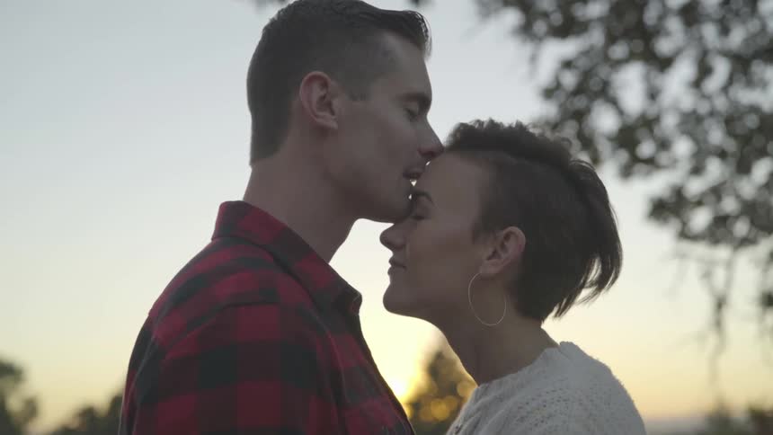 A young man and young woman stand close to each other while they kiss each other and exchange smile as they stand outside