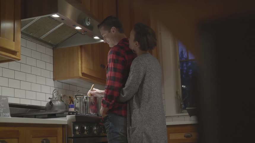 A woman hugs her husband from behind as he stirs a pot on the stove and they chat and smile