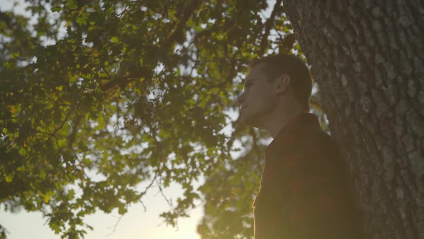 A man leans against a tree trunk outside at sunset