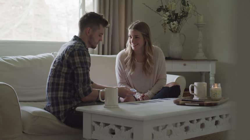 A young couple talk to one another while sitting on a white sofa on a bright day