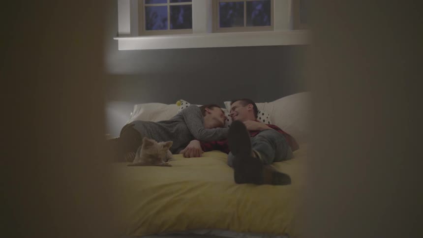 A couple cuddling on the bed with a dog playing nearby