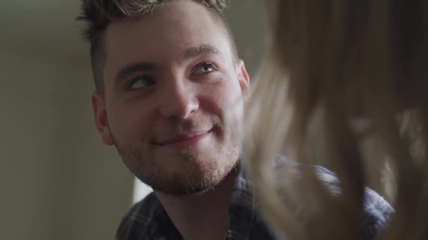 A man with a beard smiles while talking to a woman indoors