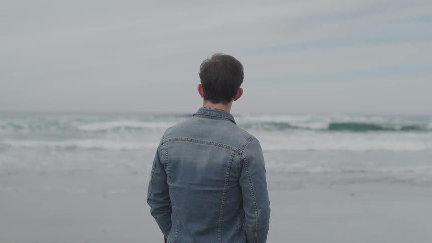 A man looks out at the ocean waves coming to shore on a gray cloudy day