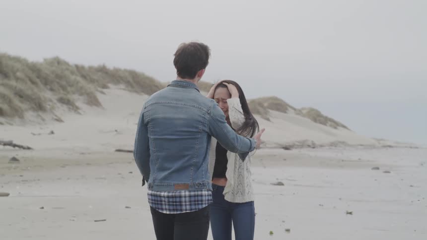 A man hugs and comforts a crying woman on a sandy beach