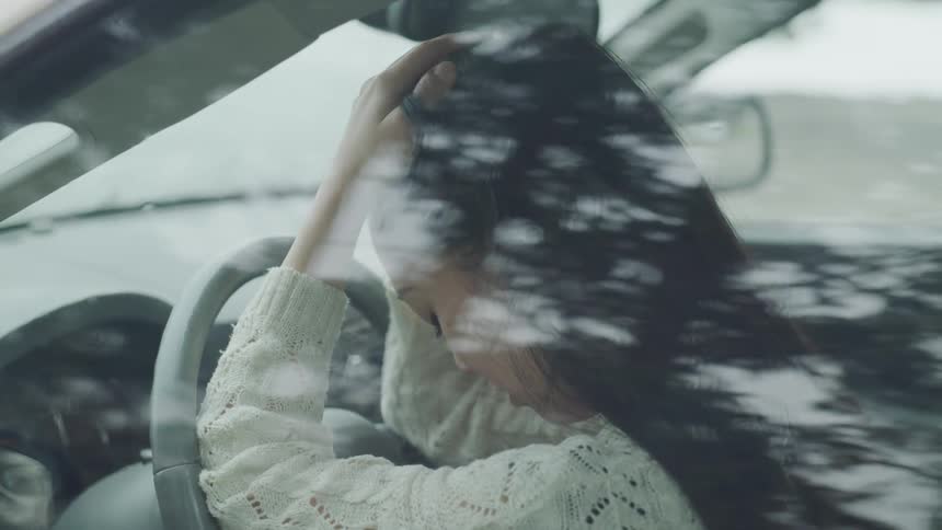 A woman sits in the driver's seat of a car and holds her head in her hands