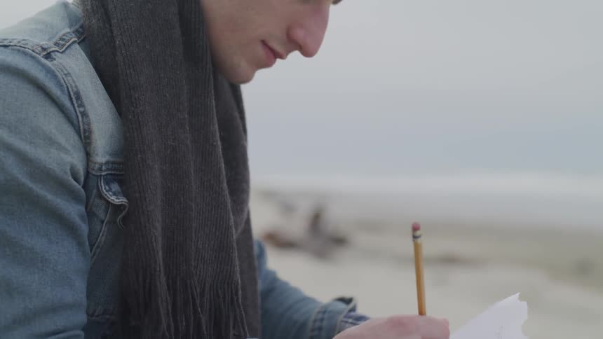 A man wearing a denim jacket and a scarf writes on a piece of paper with a pencil while sitting outside