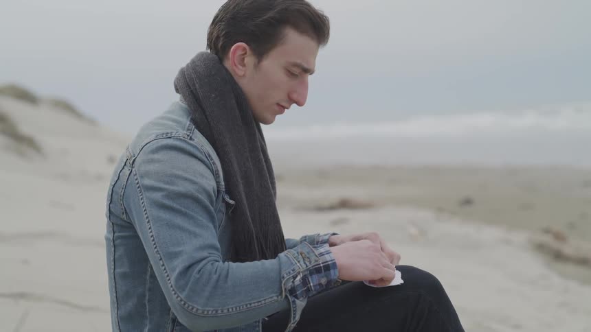 A man sits on the beach rolling up a piece of paper