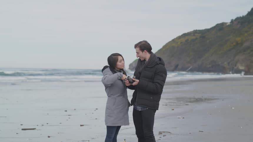 A couple stands on a beach together looking at a camera in the man's hands