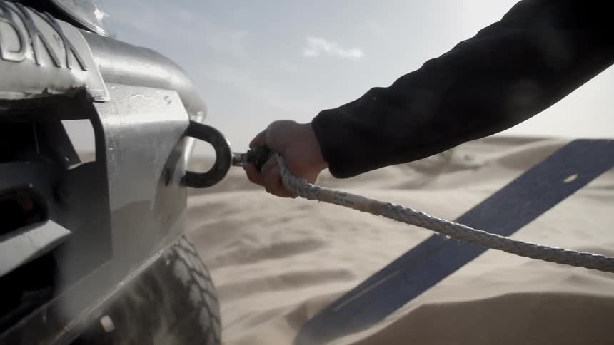 A man reaches down in the sand to pick up a large hook connected to a thick rope and attaches the hook to a connector on a buggy