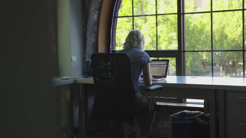 Woman sits at a desk in front of a window as she works on her laptop