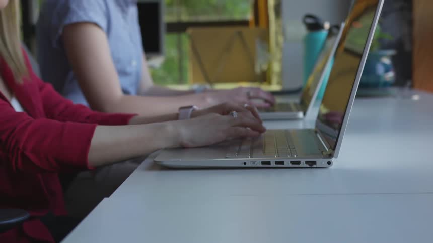 Two women sit side by side at a table and both type on laptops