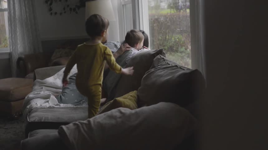 A toddler stands on a sofa and is caressed by his mother while another toddler joins them