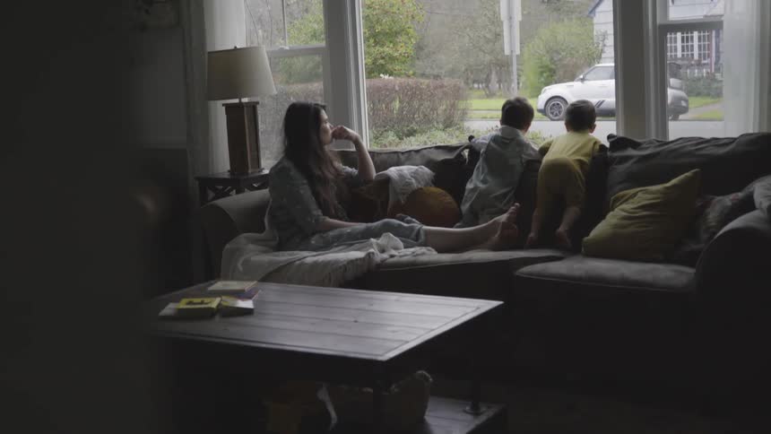 Two toddlers stand on a sofa and look out a window while their mother watches them