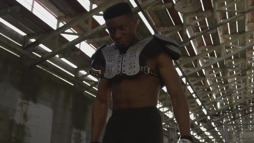 A very muscled man wearing football padding walking under the bleachers
