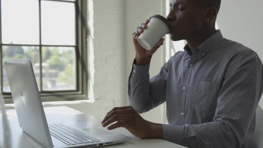 A man is typing on a laptop and drinking coffee indoors