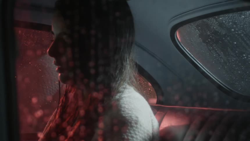 A young woman sits in the back seat of a car on a dark wet night