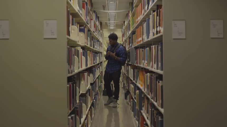 Black man wearing backpack and blue striped shirt walks and looks around in the library