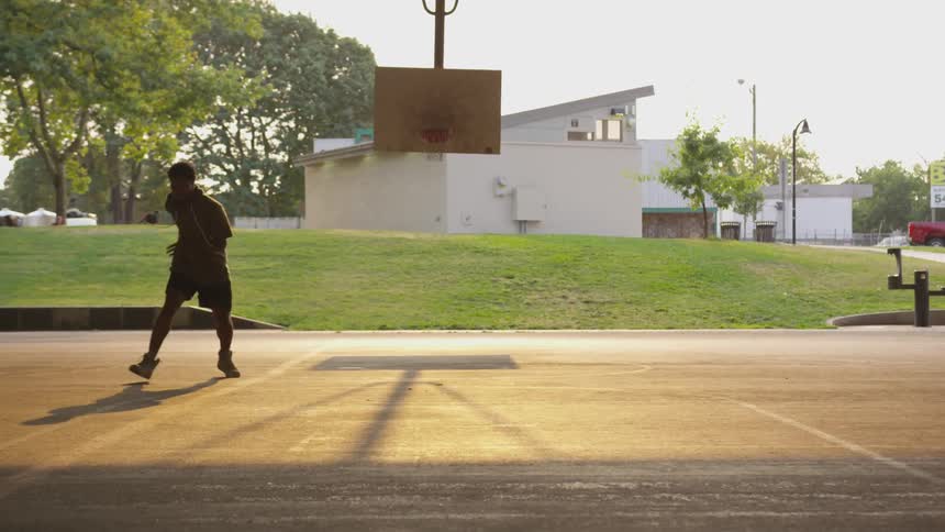 Black man wearing hoodie plays basketball on the concrete ground at dusk