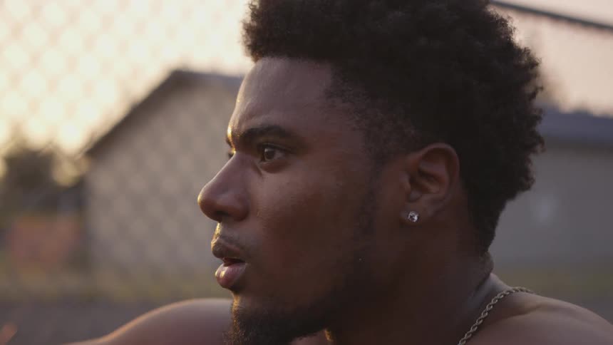 A muscled man sits to catch his breath near a chain link fence
