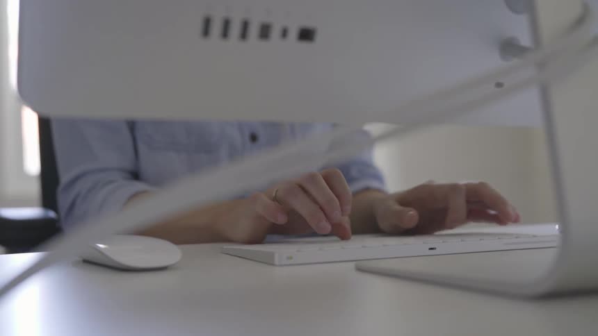 A woman's hands are typing on a keyboard her head and face are blocked by the back of the monitor