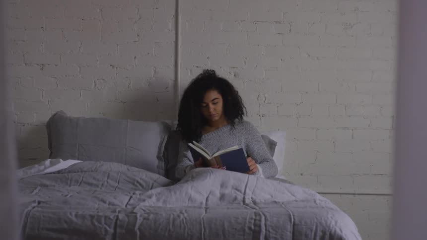 A young black woman sits in her bed while reading a book in a room with a painted brick wall