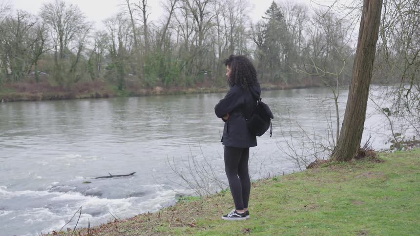A woman stands on a river bank with her arms crossed looking at the water