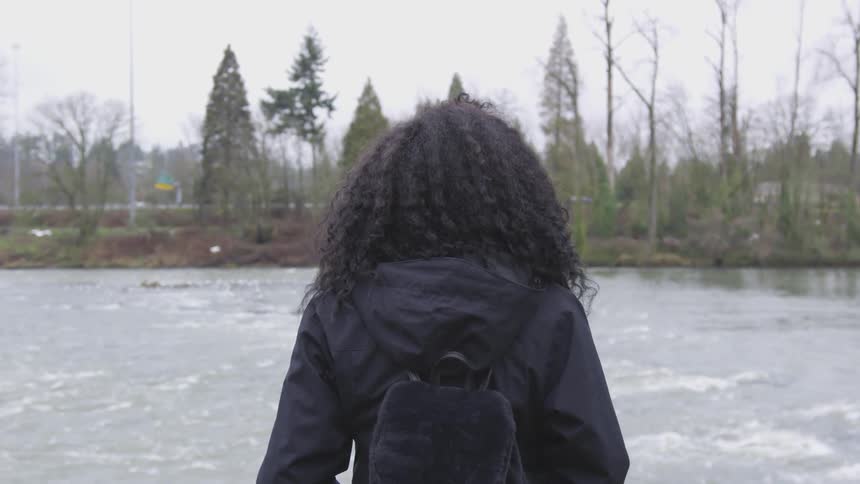 Curly hair woman wearing black backpack and coat stands in front the river on a cloudy day