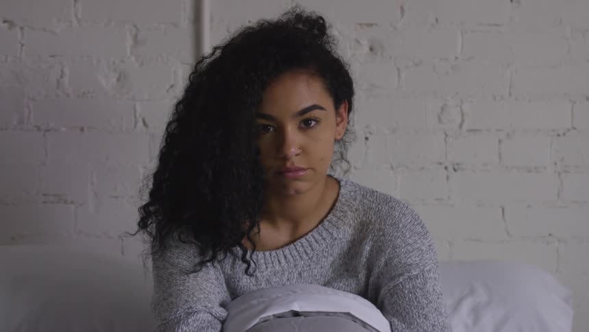 A black woman sits in bed looking down and then turns her head up