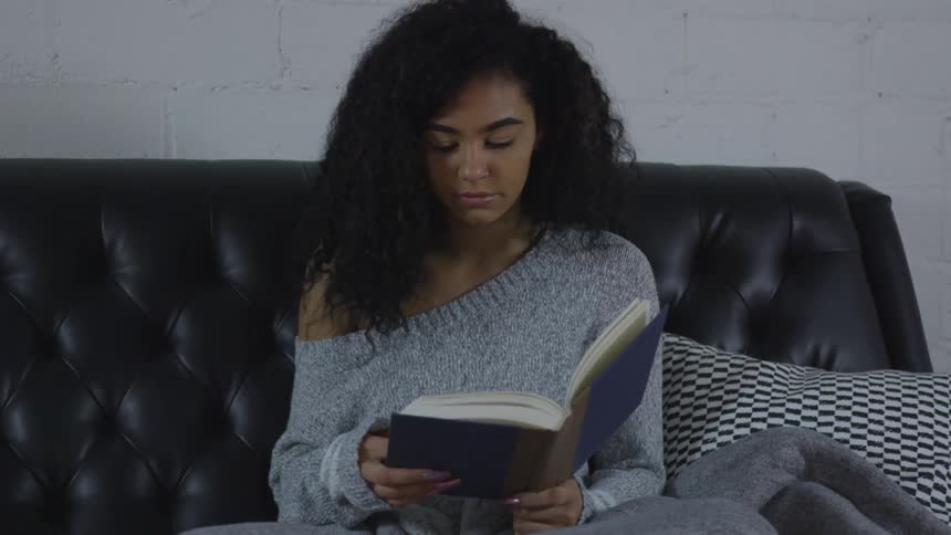 A black woman sits on a couch while reading a book