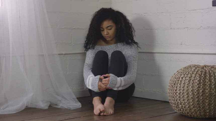 A young adult female sits in the corner of a room with her knees tucked up close to her chest and her arms wrapped around them