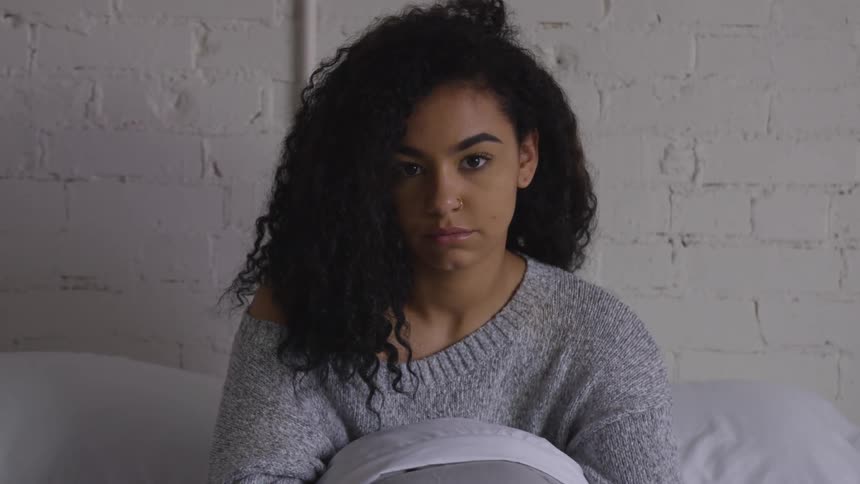 Woman sitting on the bed against a white brick wall looking at the camera