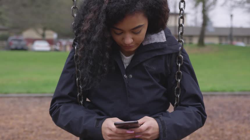 A woman sits on a swing with her arms around the chains as she texts on her phone