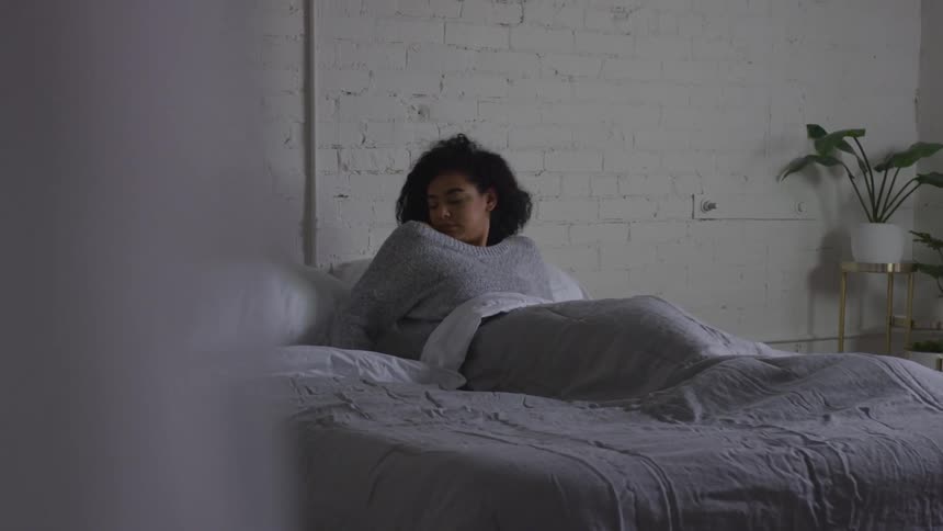 A young woman rolls over in bed before sitting up while still under the blanket