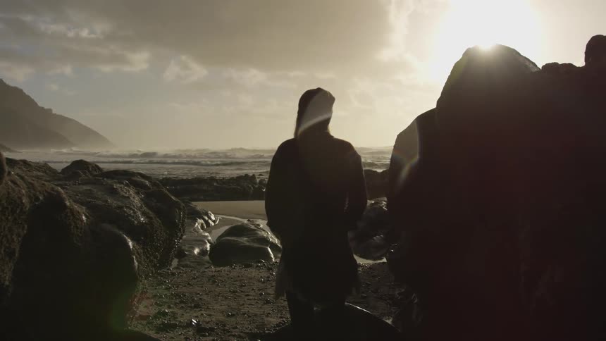 Woman wearing a hoodie stands on the rocky shores of a beach at sunset