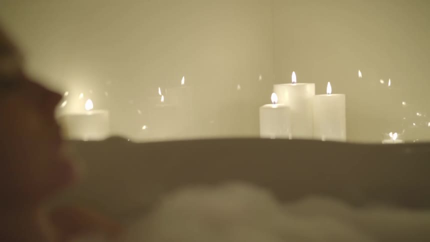 Woman soaks in bubble bath in bathroom lit by candles