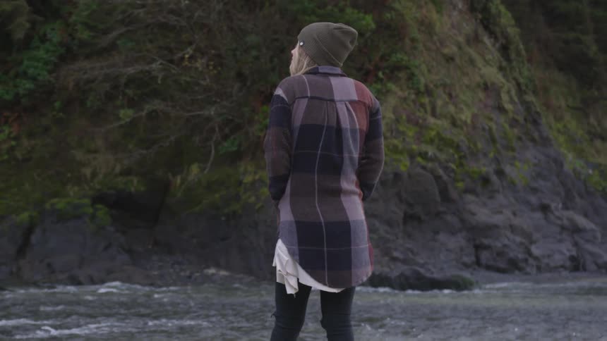 A woman standing near water and cliffs spins to take in the scenery