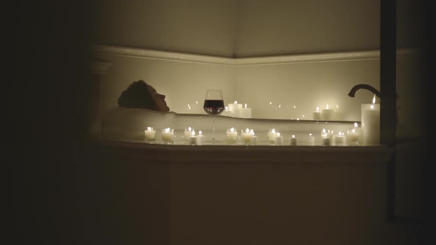 A woman lays in a bubble bath that has many candles surrounding it
