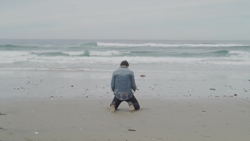 A young man standing on the beach falls to his knees