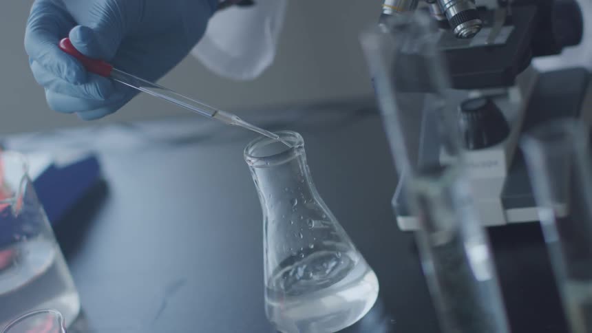 A person wearing a blue safety glove uses a dropper to drop liquid into a laboratory beaker