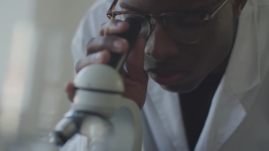 A young man wearing a lab coat is peering into a microscope
