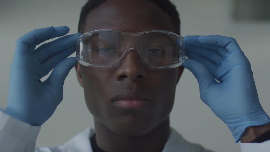 A black man wearing light blue plastic gloves adjusts his safety goggles