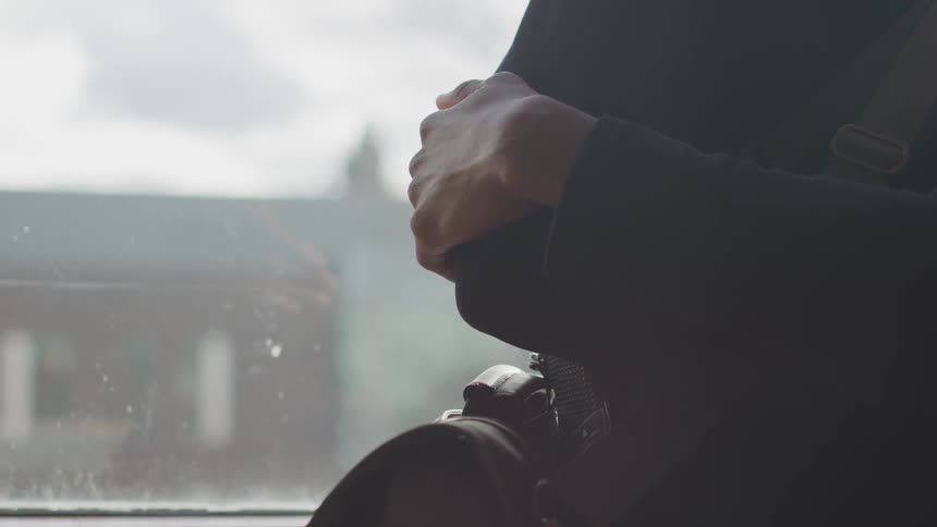 A person leans against a window with their arms crossed and a bag strap across their chest