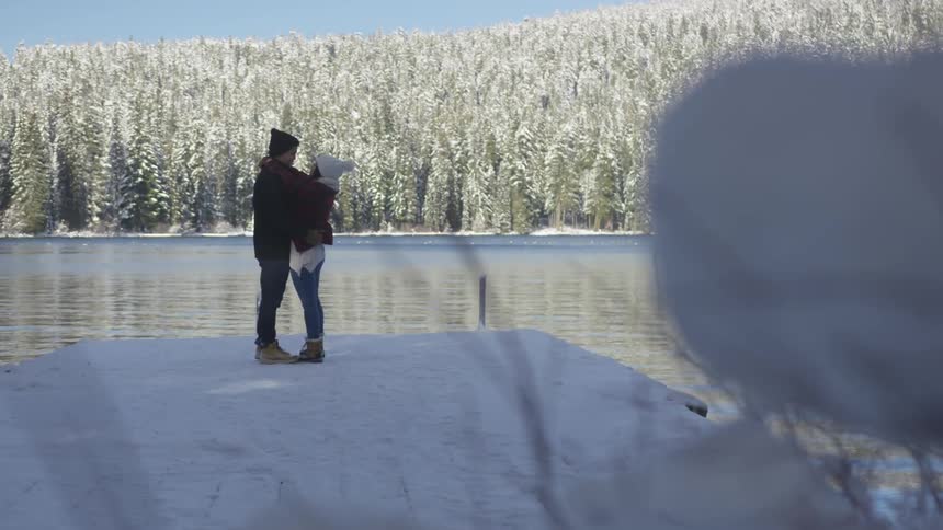 A man and a woman are holding each other and looking into each others eyes on a snowy lake dock
