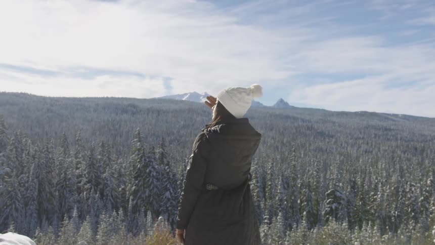 A woman stands on the edge of a cliff staring out at the snow covered trees and mountains