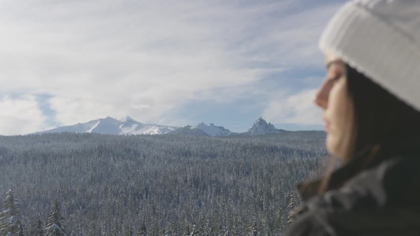 A woman stands with her eyes closed in front of a forest and mountains in the cold air