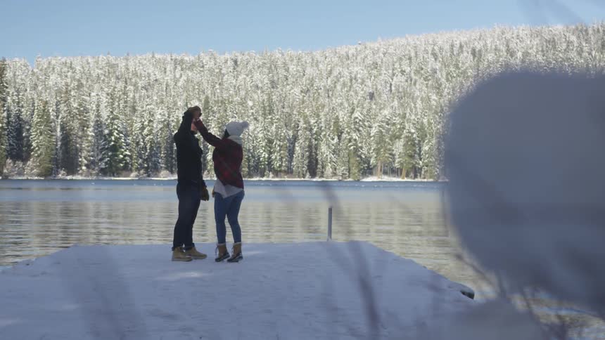 Man and woman dance on the snowy pier with the view of snowy forest by the lake on a sunny day