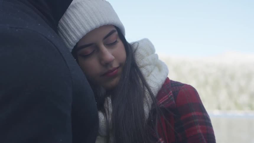 A young woman in a knit hat and heavy plaid jacket is standing peacefully snuggled next to a taller man while they are outdoors in a snow area