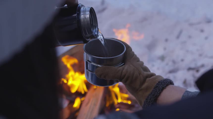 Liquid is poured from a thermos near a bonfire made on snowy ground